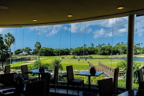 Vue depuis un espace de restauration intérieur, avec des tables et des chaises, donnant sur un terrain de golf avec un plan d’eau et des palmiers.