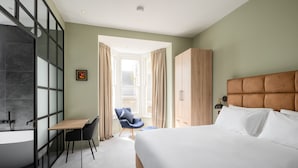 Moderni hotellihuone, jossa on suuri sänky, puinen vaatekaappi, työpöytä tuolilla ja kylpyamme.