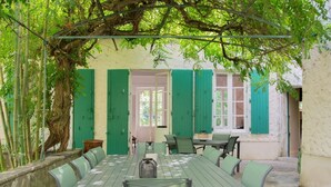 An outdoor dining area with a long table and chairs, surrounded by a white wall with green shutters, and a tree providing shade.
