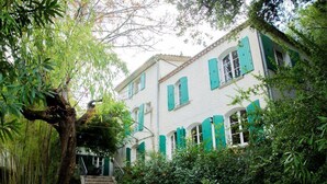 A white building with green shutters, surrounded by lush greenery and a staircase leading to the entrance.