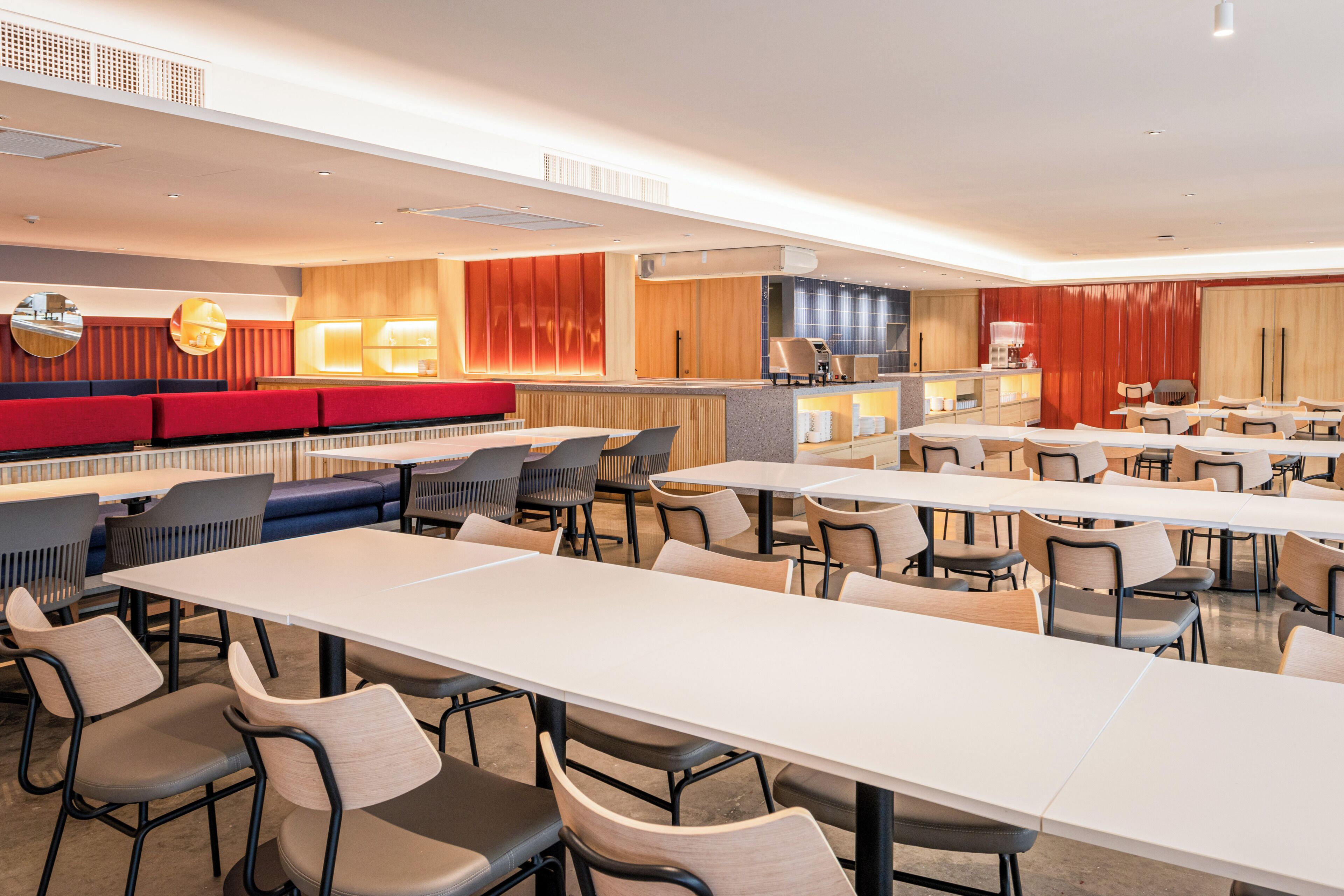 A modern dining area with multiple tables and chairs, a counter with a cash register, and a seating area with red and blue upholstered benches.