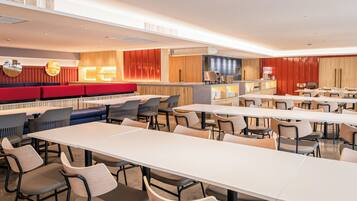 A modern dining area with multiple tables and chairs, a counter with a cash register, and a seating area with red and blue upholstered benches.