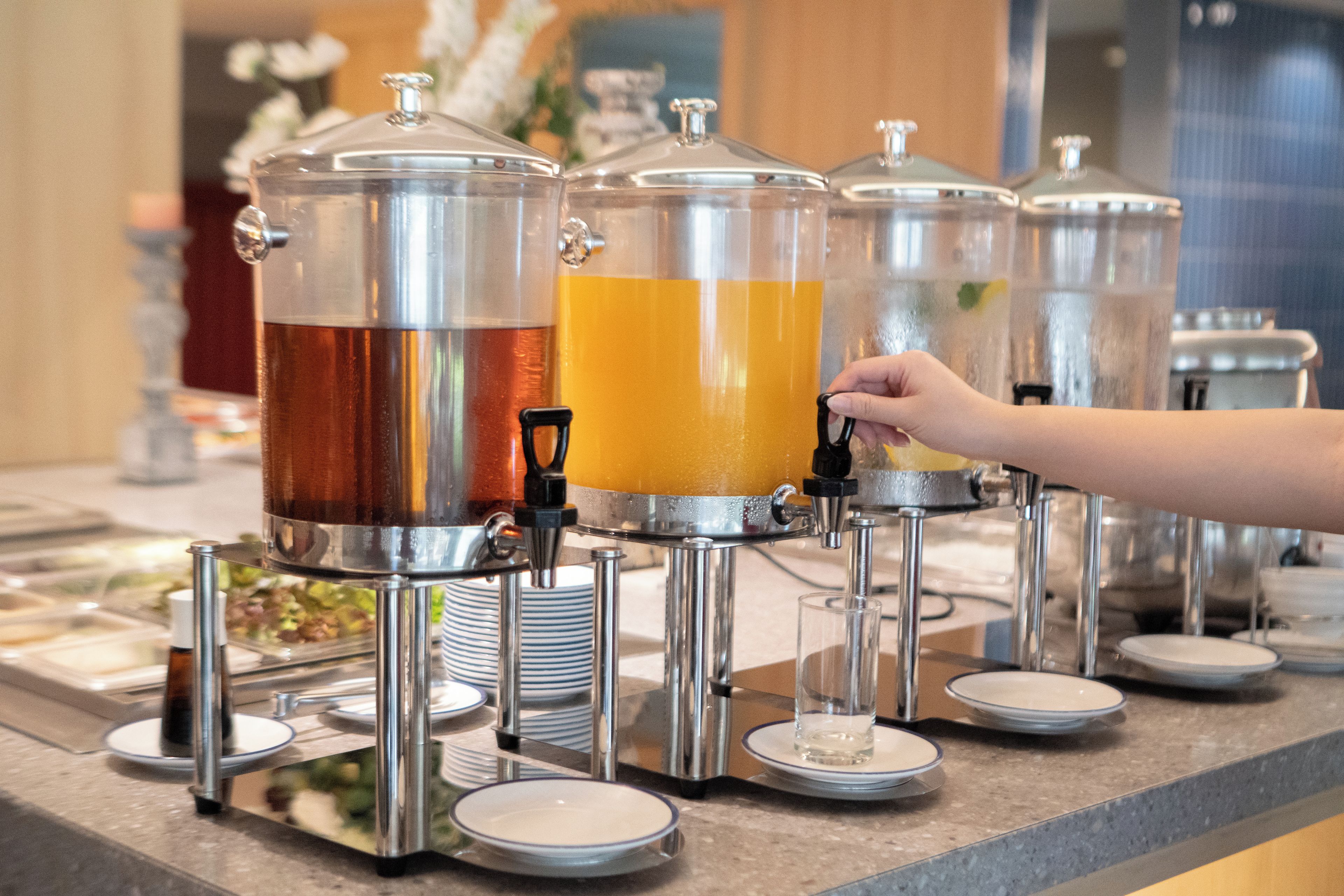 Una estación de bebidas con varios dispensadores, incluyendo uno de té y otro de jugo, y una mano alcanzando el jugo.