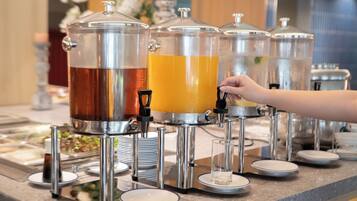 Una estación de bebidas con varios dispensadores, incluyendo uno de té y otro de jugo, y una mano alcanzando el jugo.