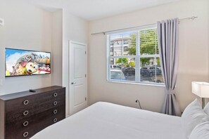 Habitación de hotel con cama, cómoda, televisor en la pared, ventana con cortinas y vista a la calle.