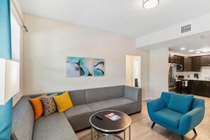 A modern living room with a grey sectional sofa, a blue armchair, and a round coffee table. - Resort Condo: 3BR w Balcony & Parking near Disney! (Orlando)