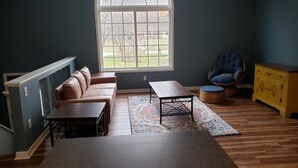 Una habitación con un amplio ventanal arqueado, un sofá de cuero marrón, una mesa de centro de madera, una silla de mimbre y un armario amarillo.