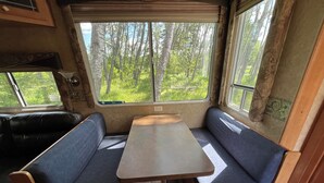 A compact RV interior with a central table and two bench seats, large windows offering a view of a forested area.