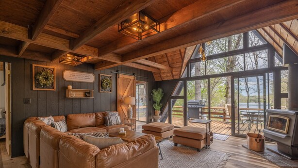 A cozy cabin interior with a leather sofa, wooden flooring, a fireplace, and large windows offering a view of the outdoors. - Happy Moments Chalet by Innsbrook Vacations! (Innsbrook)