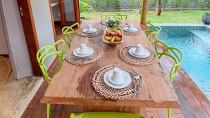 Une table en bois, dressée pour quatre personnes, avec des chaises assorties, un panier de fruits et une vue sur une piscine et des palmiers.