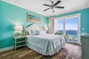 Ein Schlafzimmer mit Bett, Nachttischen, Deckenventilator, einem Gemälde und einem Balkon mit Meerblick.