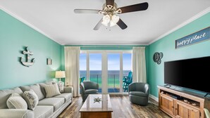 Een woonkamer met een bank, stoelen, een salontafel, een televisie en een balkon met uitzicht op de oceaan.