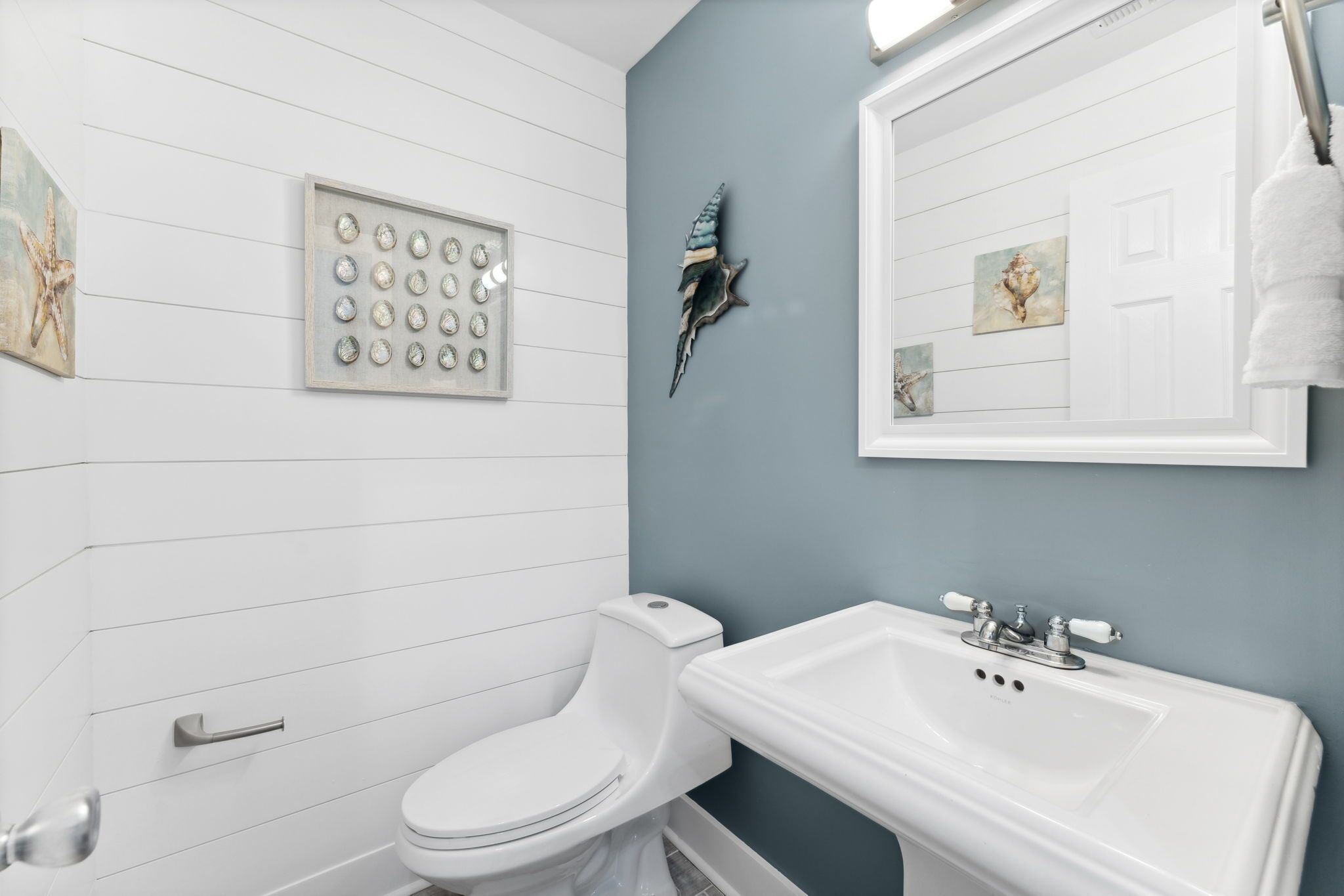 Une salle de bain avec des toilettes blanches, un lavabo et des éléments décoratifs sur les murs, notamment une coquille et des tableaux encadrés.