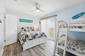 Une chambre à coucher comprenant un lit, un lit superposé, un ventilateur de plafond, une porte et une fenêtre avec des stores.