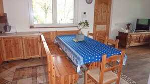 Un área de comedor con una mesa de madera y bancos, un mantel azul, una planta en maceta y un televisor sobre un mueble.