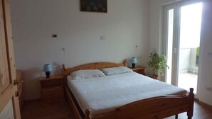Un dormitorio con una cama de madera, dos mesitas de noche con lámparas, una planta en maceta y un ventanal corredizo a un balcón.