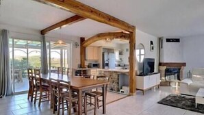 A spacious living area with a wooden dining table and chairs, a kitchen in the background, and a fireplace. - The Vault of the Stars overlooks the Monts d’Ardèche (Genestelle)