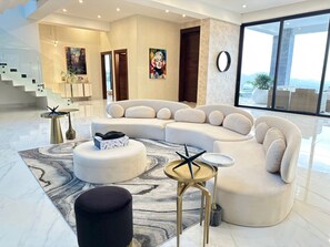 A modern living room with a curved white sofa, a round ottoman, and a small round table. There is a staircase in the background leading to an upper level.