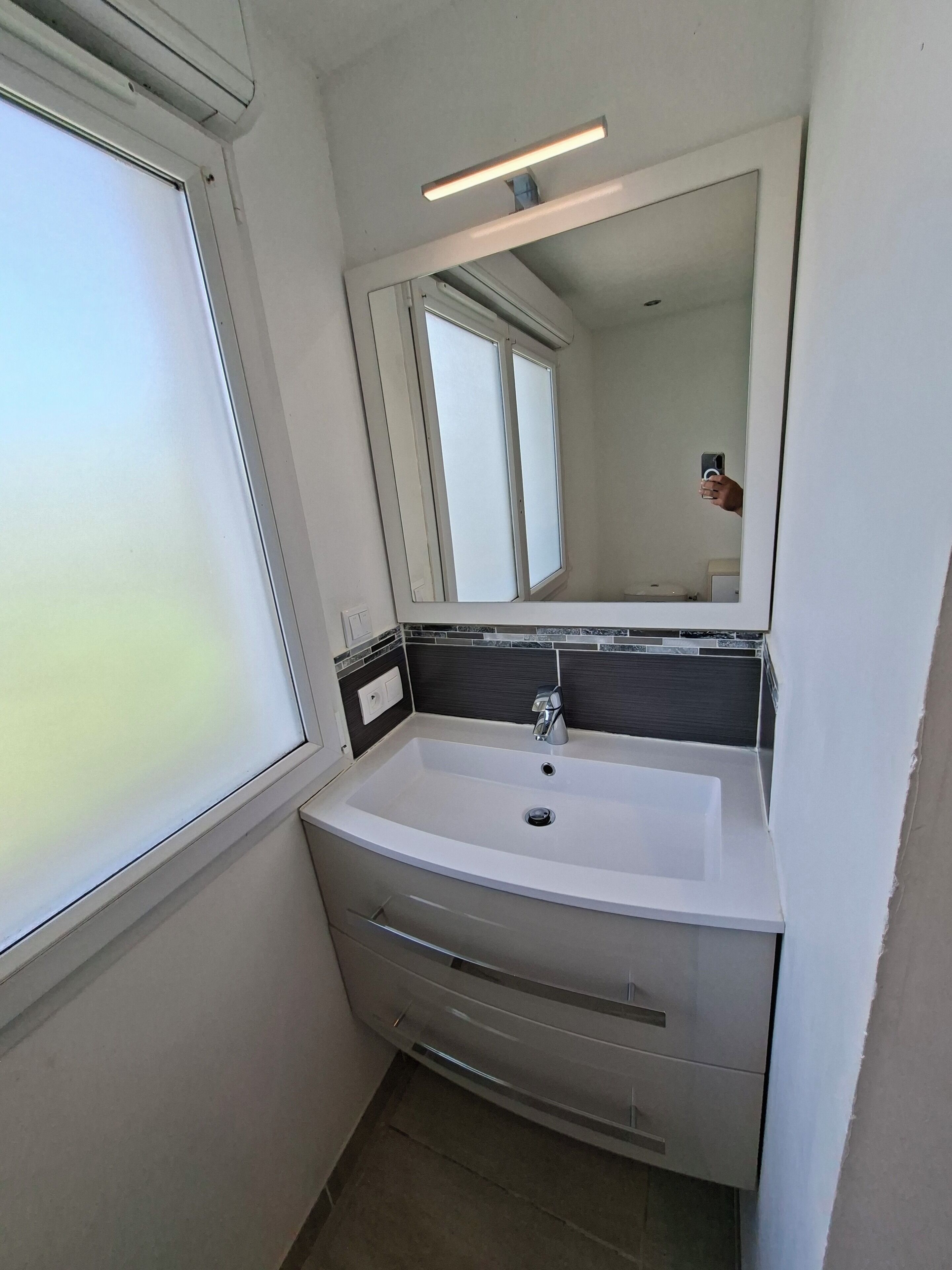 Une salle de bain avec un lavabo blanc, un miroir et une fenêtre.