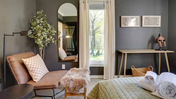 A modern hotel room with a brown leather sofa, a fluffy stool, a bed with a quilted green bedspread, a wooden desk, and a large mirror. - Mid-Century Modern Fayetteville Home Close to Town (Fayetteville)