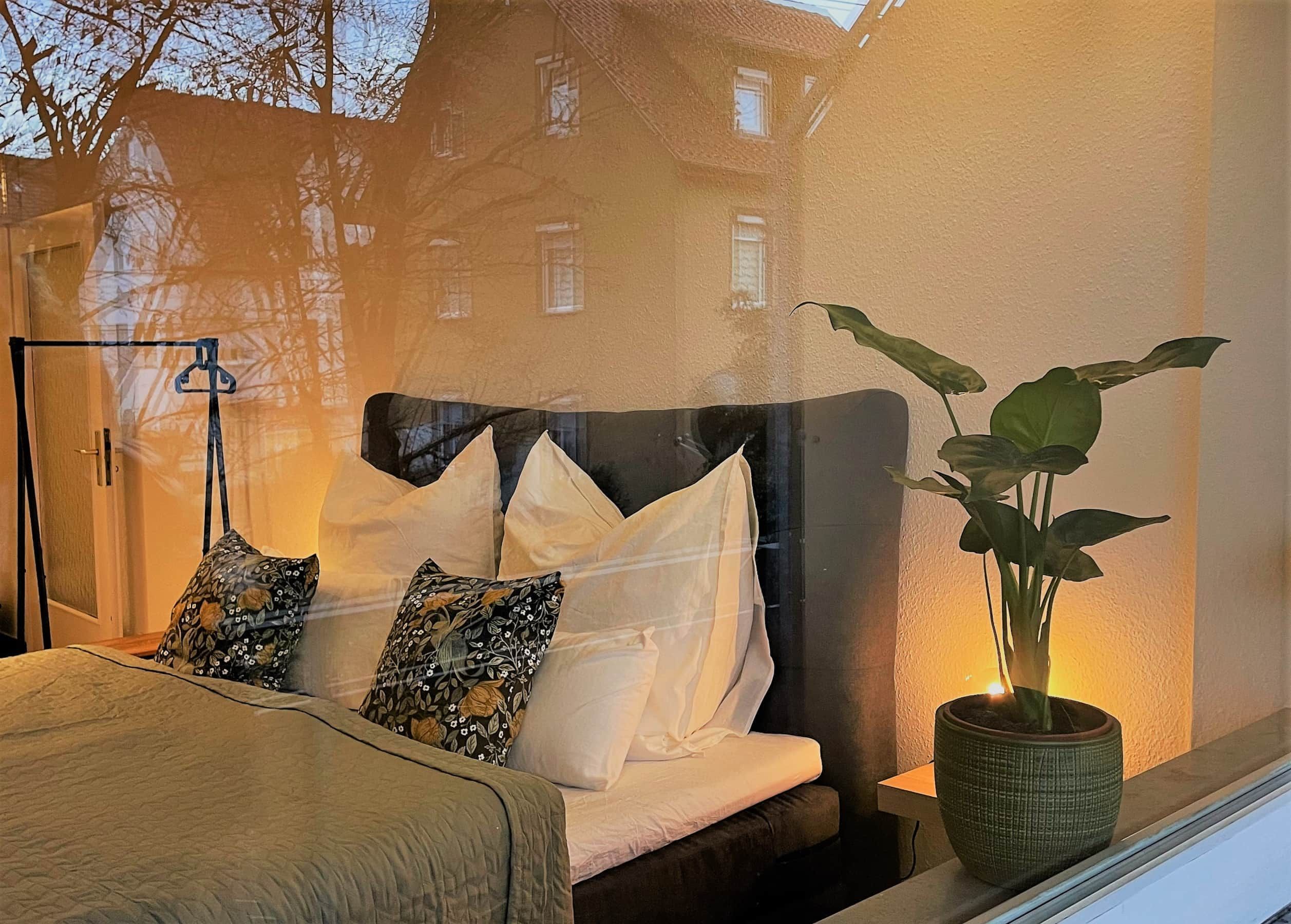 A bedroom with a bed, pillows, a bedside table with a plant, and a window with a view of buildings.