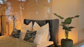 A bedroom with a bed, pillows, a bedside table with a plant, and a window with a view of buildings.