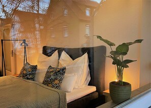 A bedroom with a bed, pillows, a bedside table with a plant, and a window with a view of buildings. - Studio apartment with parking (Esslingen am Neckar)