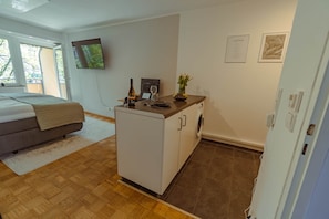 A compact hotel room with a bed, a small kitchenette, a washing machine, and a TV mounted on the wall.