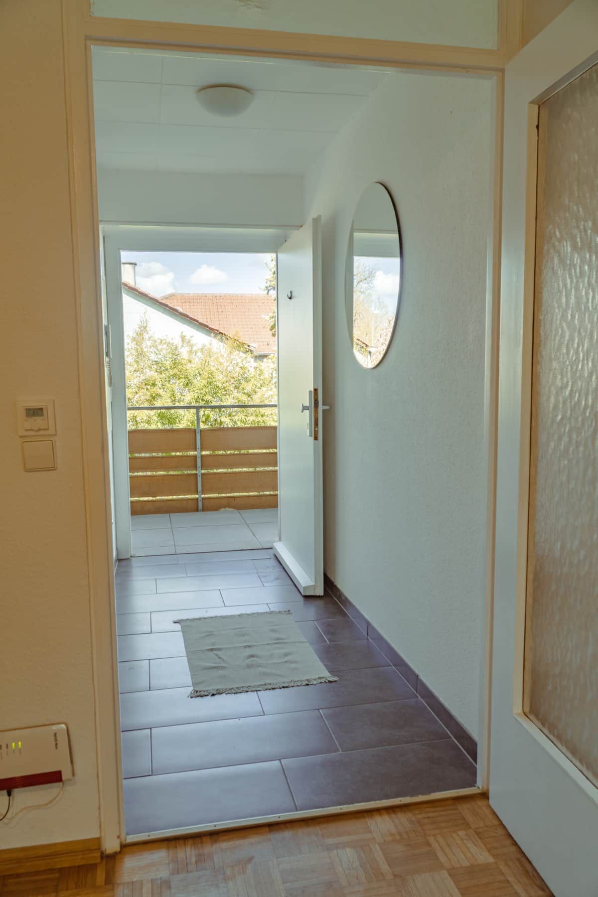 Ein Flur mit Holzboden, einem kleinen Teppich und einer Tür, die auf einen Balkon mit Blick auf Bäume und ein Gebäude führt.