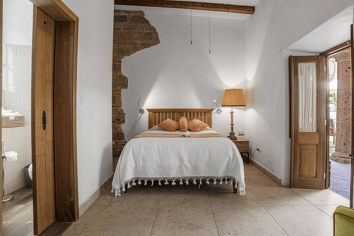 Une chambre à coucher avec une tête de lit en bois, un lit recouvert de draps blancs, une table de chevet avec une lampe, et une vue sur l’extérieur par une porte.