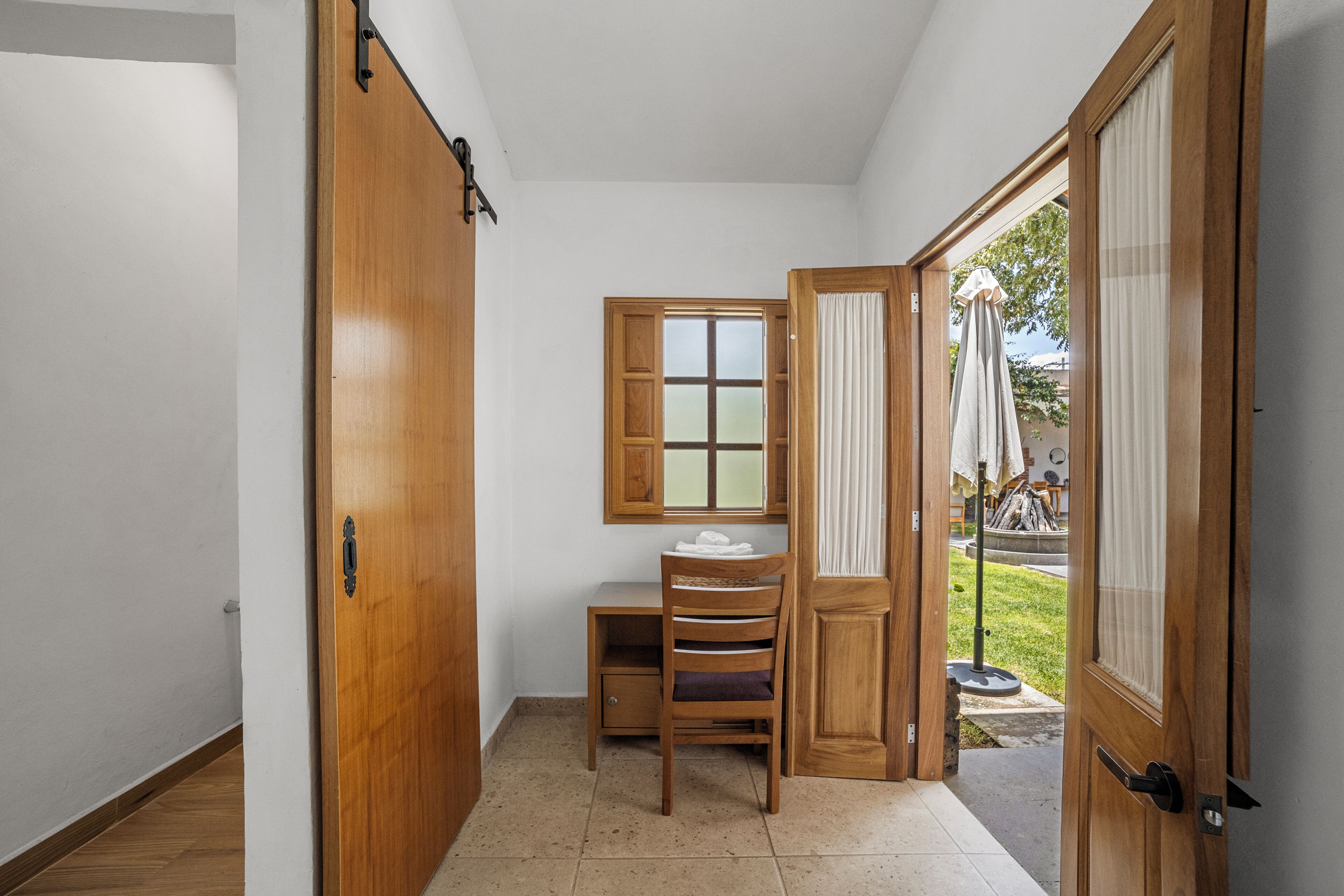 A narrow hallway with a wooden door, a window with frosted glass, a wooden chair, and a small table with a towel.
