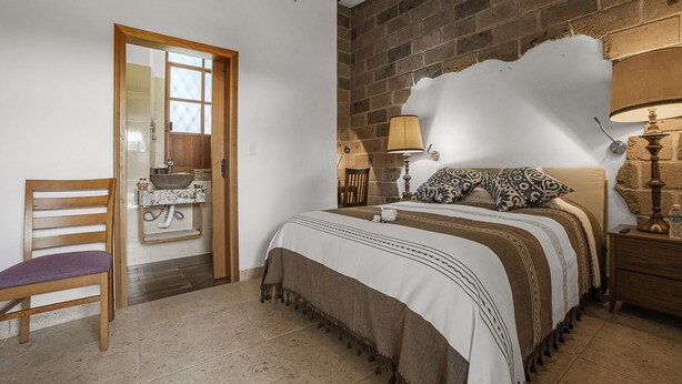 A bedroom with a bed, bedside lamps, a chair, a wooden door, and a stone wall.