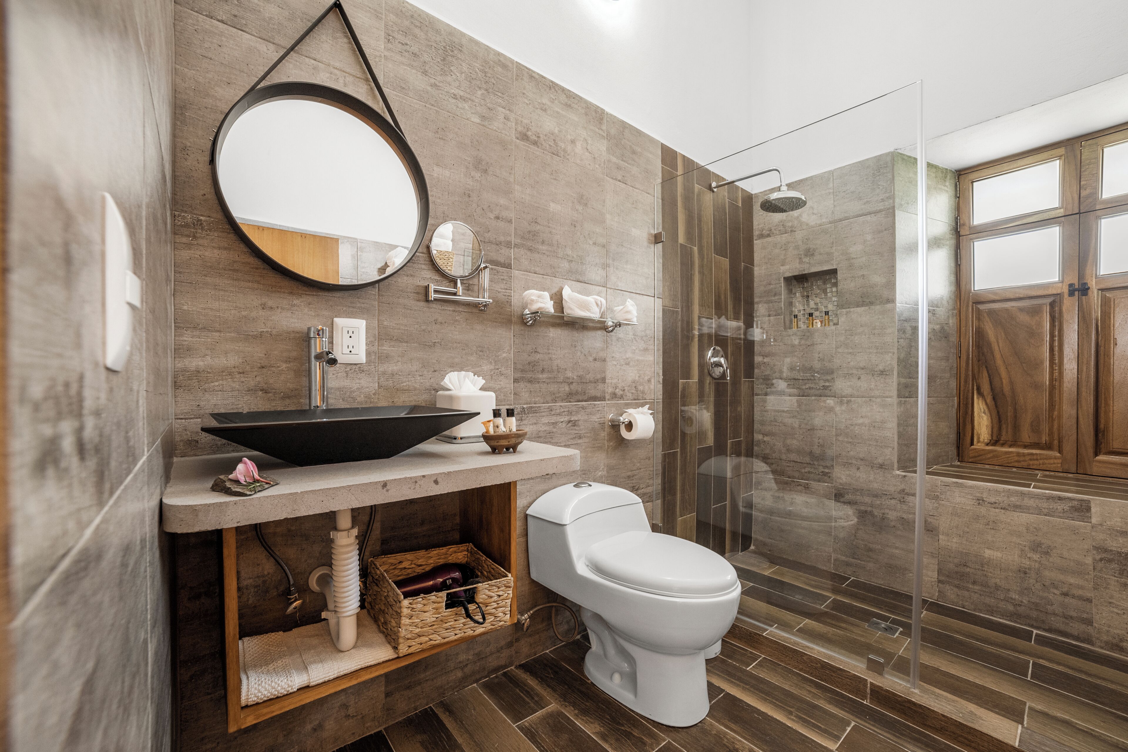 Un baño moderno con un espejo grande, un lavabo con lavamanos negro, un inodoro y una zona de ducha con mampara de vidrio.
