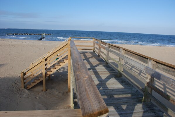 Une promenade en bois mène à une plage de sable, avec une digue rocheuse en arrière-plan.