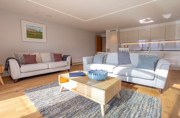 A modern living room with two sofas, a glass coffee table, and a kitchen in the background. - Square Green (No 16) | With Balcony (St Andrews)