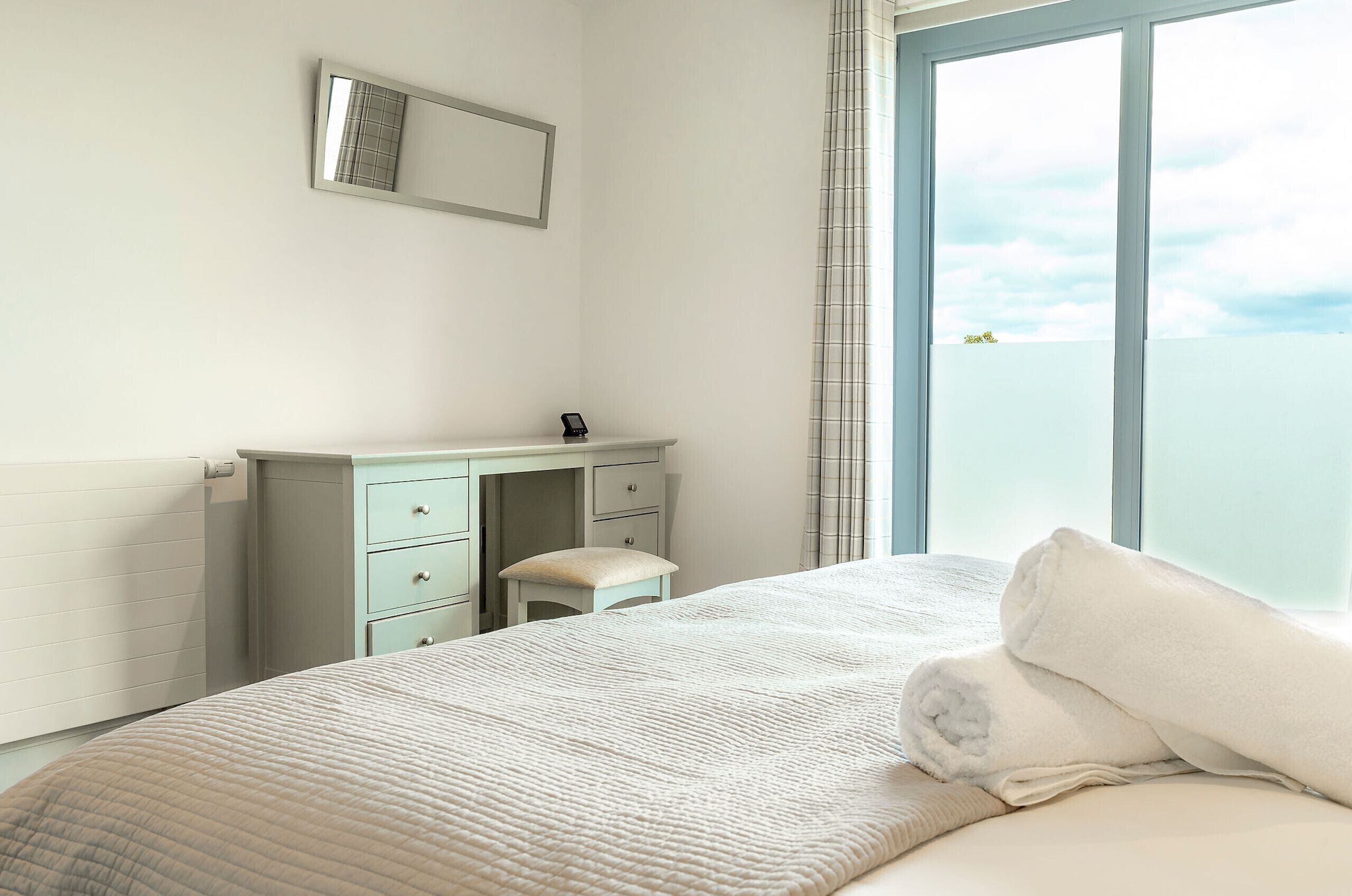 A bedroom with a bed, a dresser, a mirror, and a view of the sky through a window.