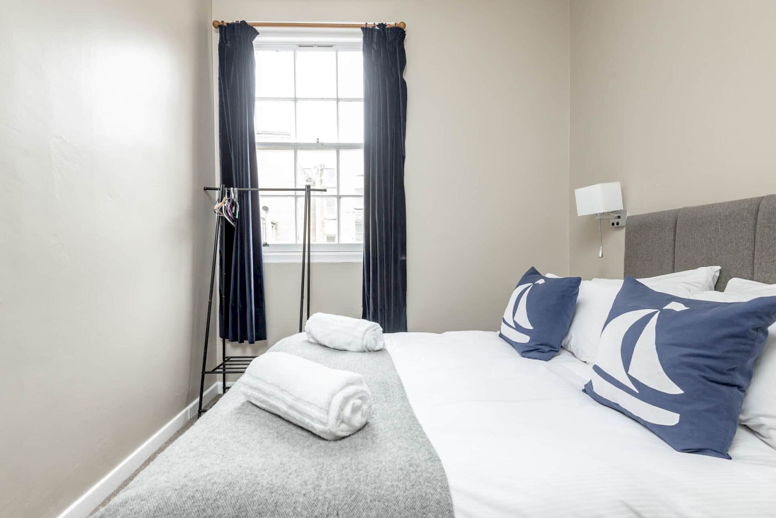 A hotel room with a bed, a window with curtains, a towel rack, and a wall-mounted lamp.