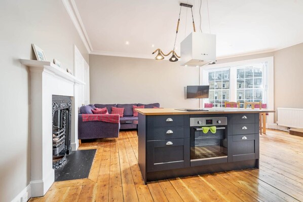 A modern kitchen with a central island, wooden flooring, a fireplace, and a sofa in the adjacent living area.