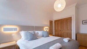 A neatly made bed with white and gray bedding, two bedside lamps, and a wooden wardrobe. - 30C The Scores (St Andrews)