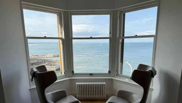 A corner room with a bay window overlooking the sea, two modern chairs, and a radiator. - 30C The Scores (St Andrews)