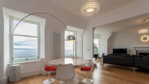 A modern living room with a round table and white chairs, a large window with a view of the sea, and a black sofa. - 30C The Scores (St Andrews)