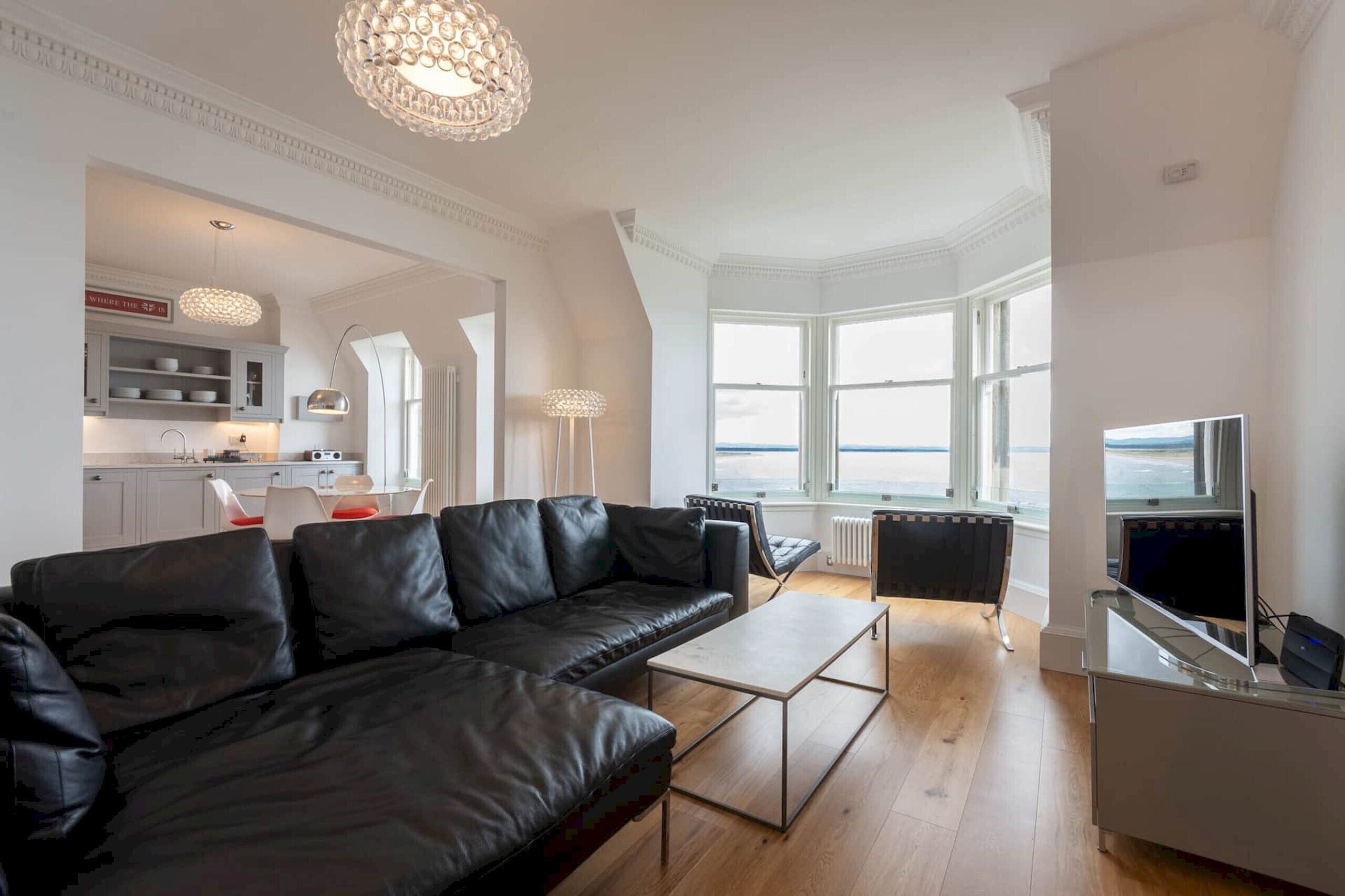 A modern living room with a black leather sofa, a coffee table, and a TV stand. There is a kitchen area in the background with white cabinets and a dining table. Large windows provide a view of the outdoors.