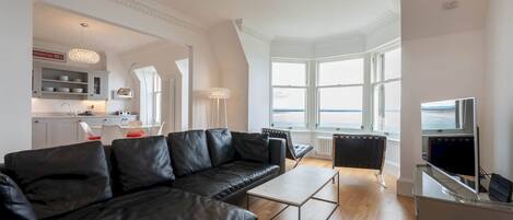 A modern living room with a black leather sofa, a coffee table, and a TV stand. There is a kitchen area in the background with white cabinets and a dining table. Large windows provide a view of the outdoors.