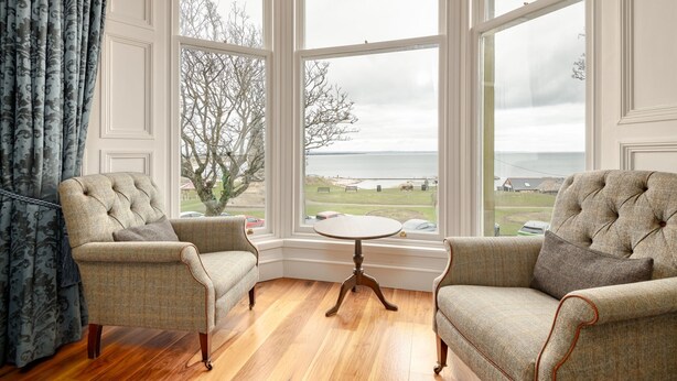 A corner room with two armchairs, a small round table, and large windows offering a view of the sea and a golf course. - Gillespie Apartments | Golfer's Dream (St Andrews)