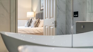 A modern bathroom with a bathtub, a shower area, and a visible part of a bedroom with a bed and a nightstand. - Gillespie Apartments | Golfer's Dream (St Andrews)