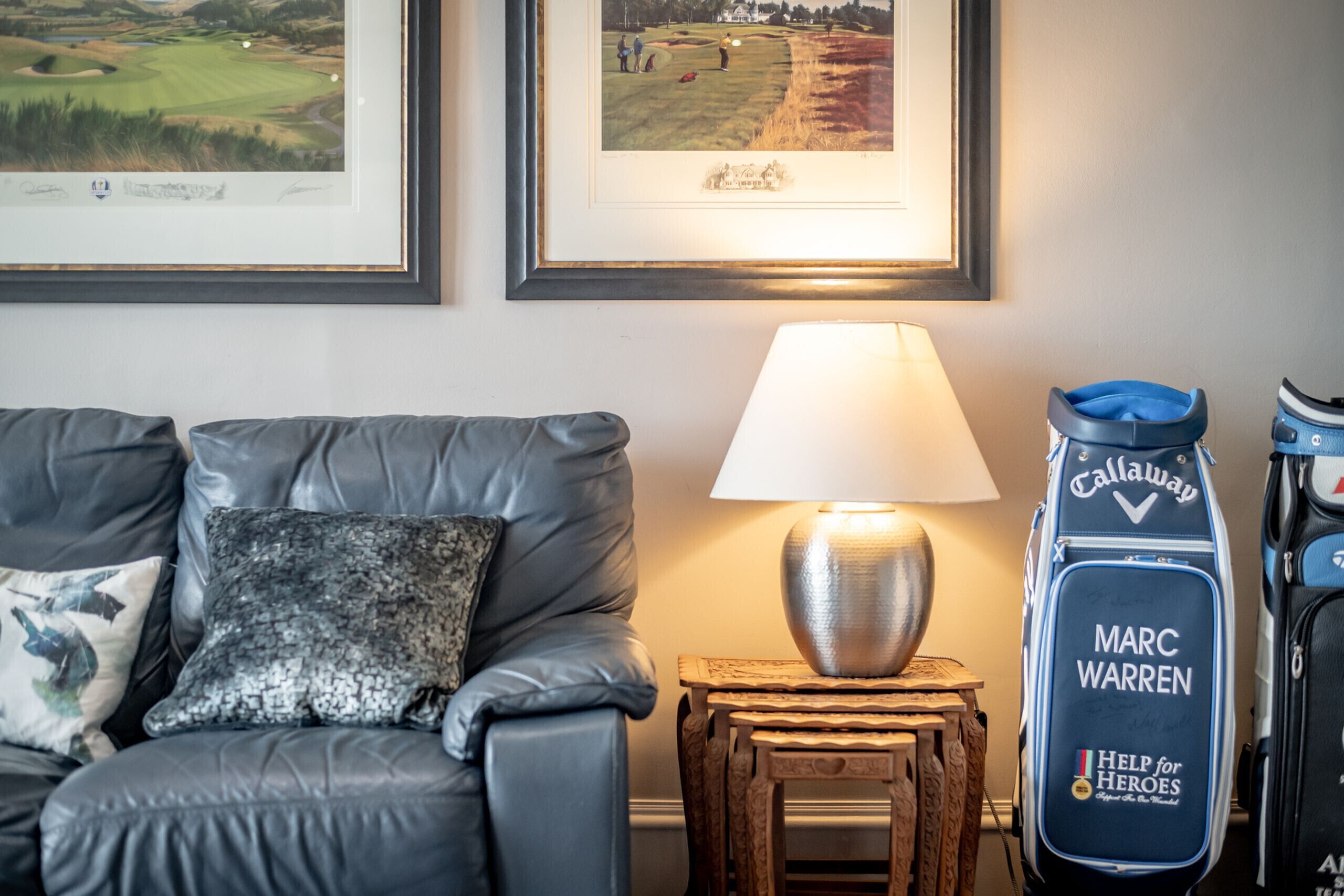 A room with a leather couch, a lamp, framed golf pictures, and a golf bag.