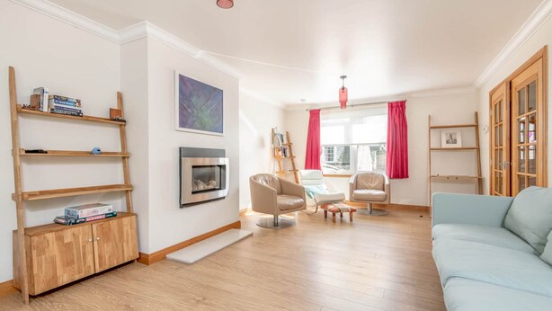 A spacious living room with a light blue sofa, wooden shelves, a fireplace, and a large window with red curtains. - Dempster Lodge (St Andrews)