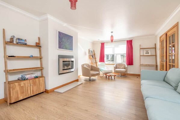 A spacious living room with a light blue sofa, wooden shelves, a fireplace, and a large window with red curtains. - Dempster Lodge (St Andrews)