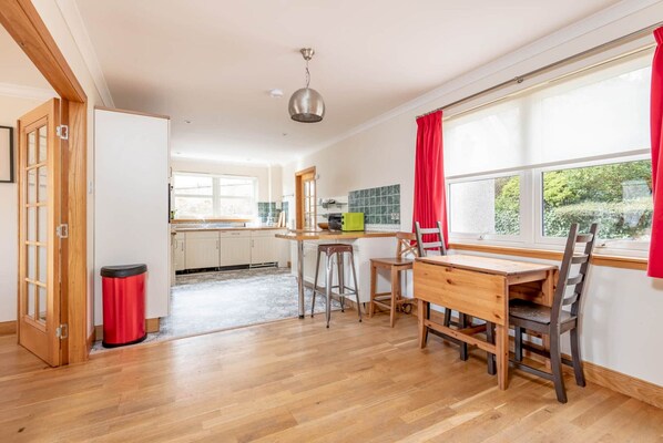 A modern kitchen with wooden flooring, a dining area with a wooden table and chairs, a red trash bin, and a window with red curtains. - Dempster Lodge (St Andrews)
