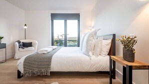 Une chambre à coucher avec un lit, une chaise, une table de chevet ornée d’un vase et une fenêtre donnant sur un paysage agréable.